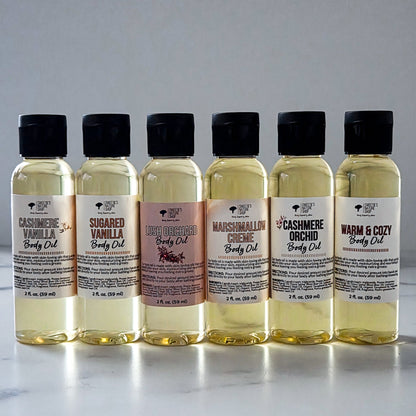 Six small clear bottles of body oil arranged in a row on a light marble-like surface against a soft gray background. The bottles have black caps and cream labels with different scent names: Cashmere Vanilla, Sugared Vanilla, Lush Orchard, Marshmallow Creme, Cashmere Orchid, and Warm & Cozy.